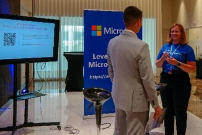 Two people communicating at an MS Learn Booth. The room is dimly light, and there is a TV and a banner beside it.