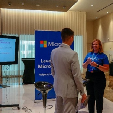 Two people communicating at an MS Learn Booth. The room is dimly light, and there is a TV and a banner beside it.