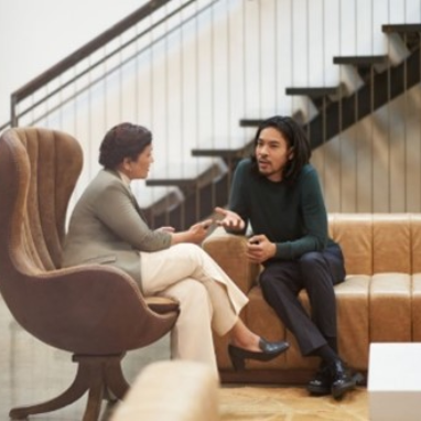 Two people, collaborating in an open space, with couches and stairs in the background.